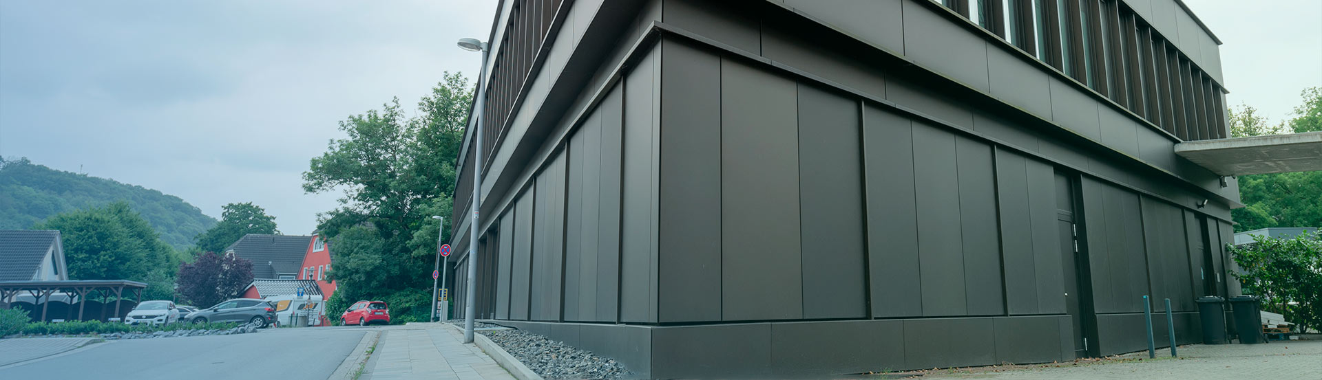 Ein modernes, dunkelgraues Gebäude mit großen Paneelen auf einem Eckgrundstück, neben einer ruhigen Straße mit geparkten Autos und Häusern, mit Grün und Bäumen im Hintergrund unter einem bewölkten Himmel.